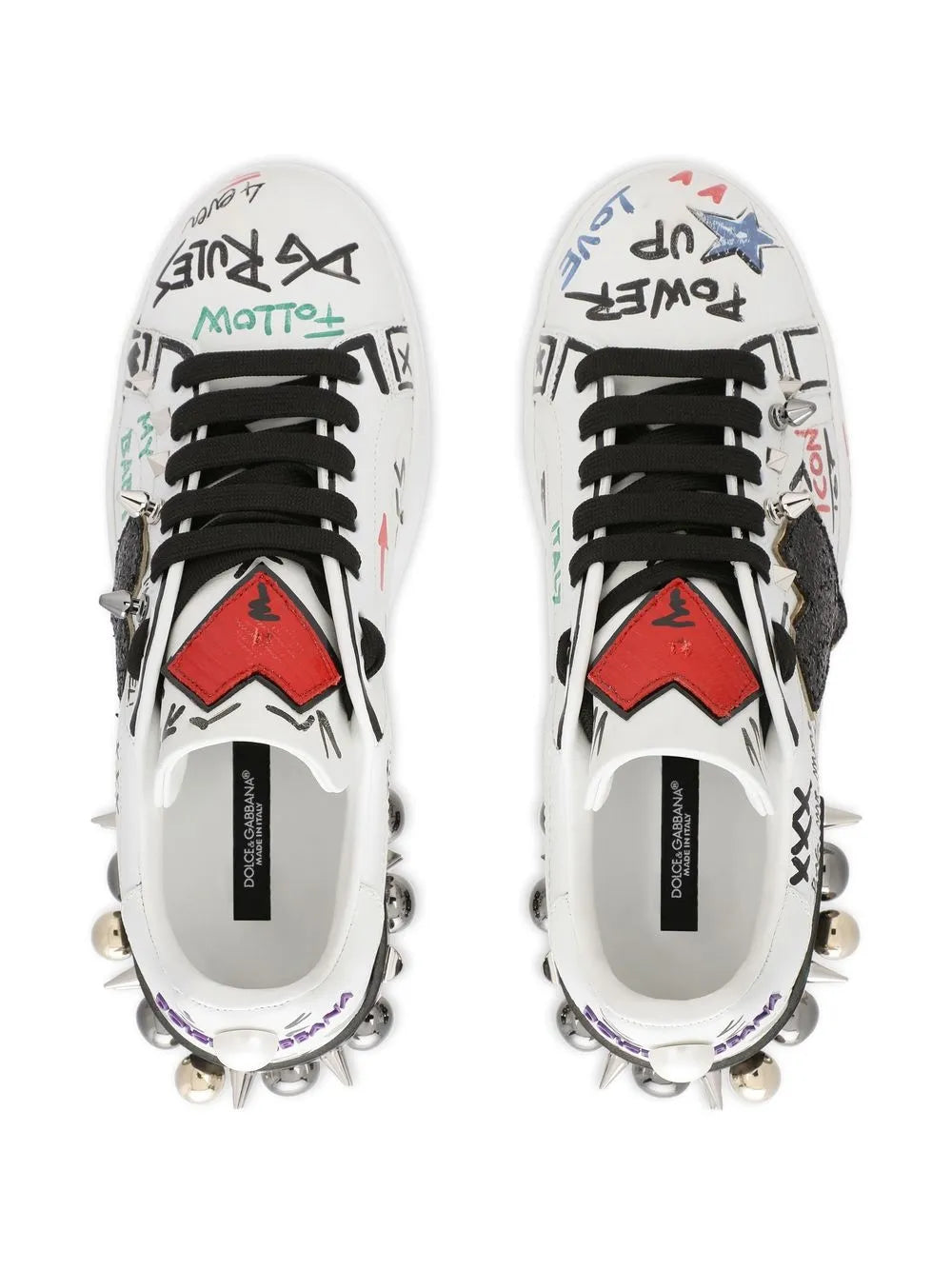 White leather sneakers with graphic tongue patches, black laces, and silver studs