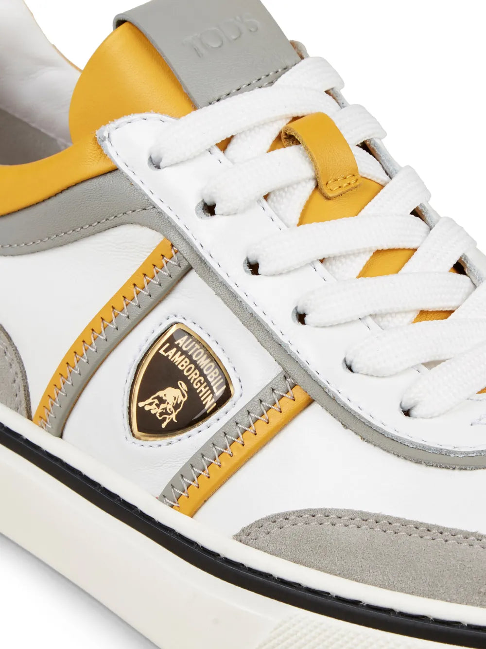White low-top sneaker close-up: gold logo patch, yellow/gray side panel, lace-up.