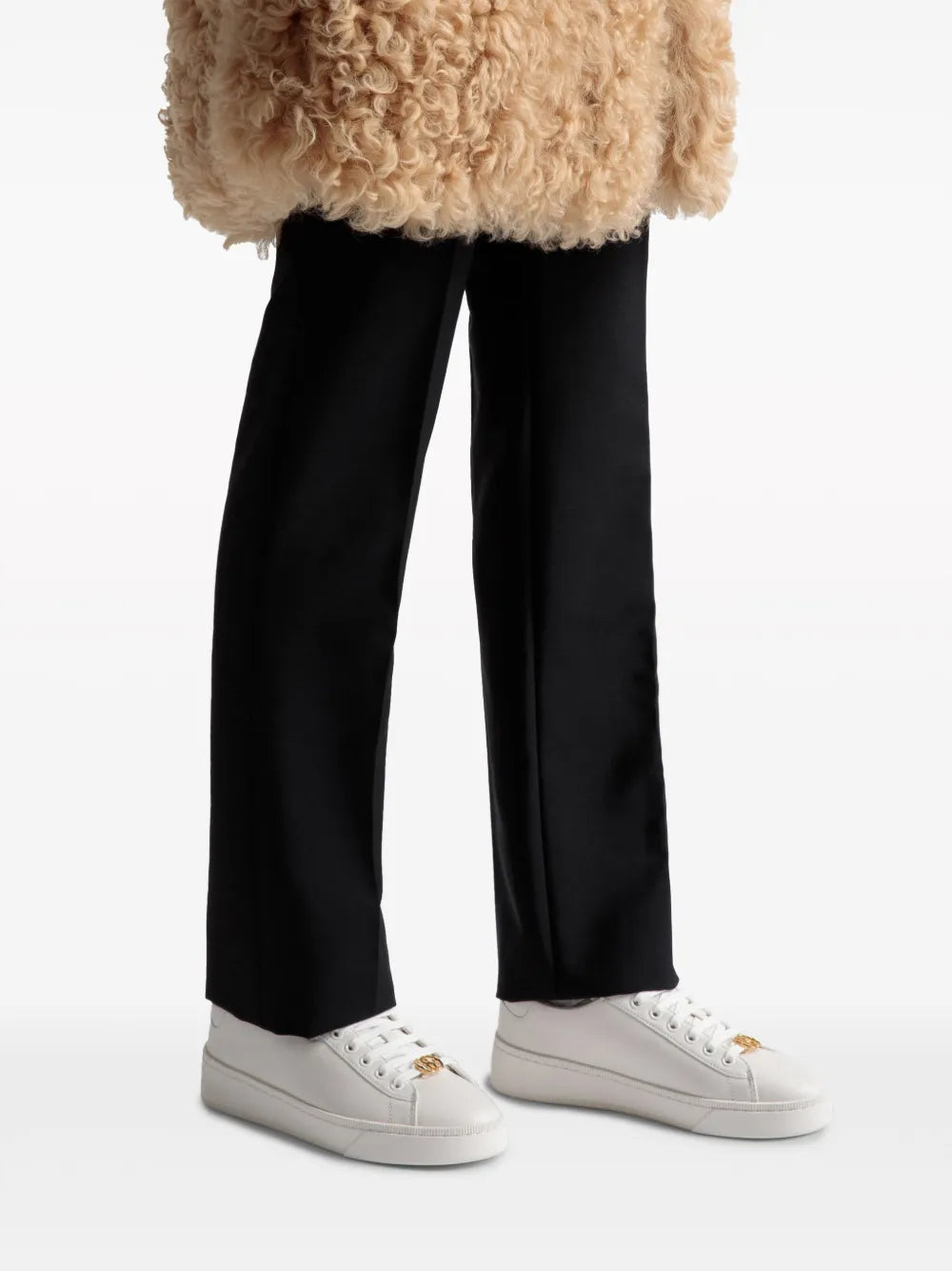 White leather sneakers on feet with gold emblem, worn with black pants