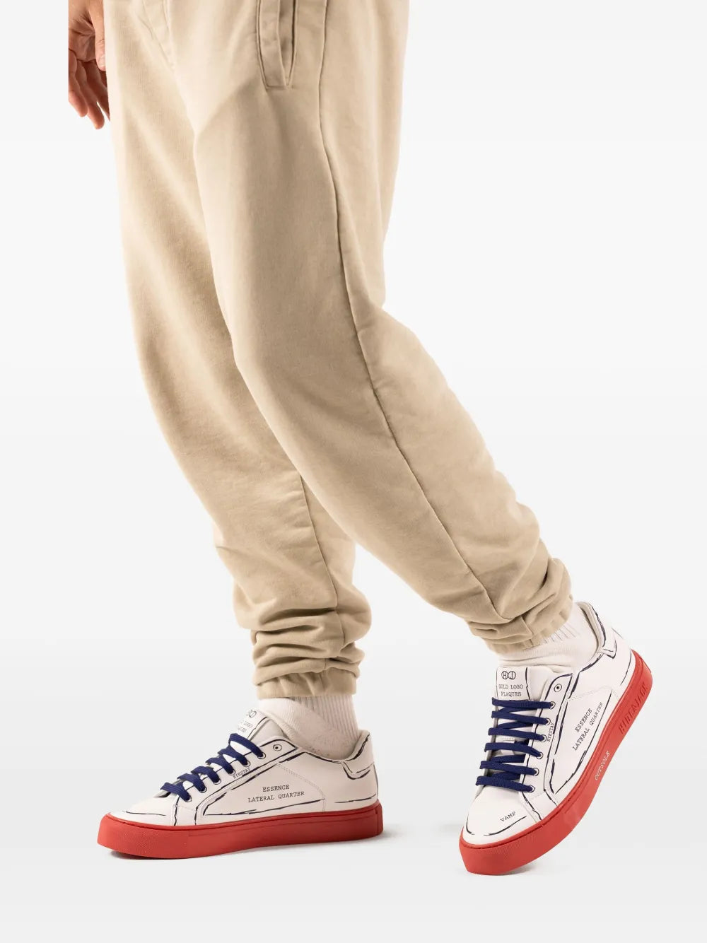 White low-top sneakers with blue laces and red outsole, worn with beige joggers