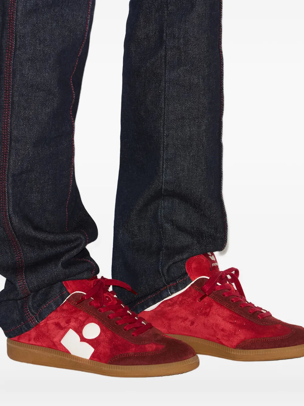 Red suede sneakers with white logo and gum sole under dark jeans, close angle.
