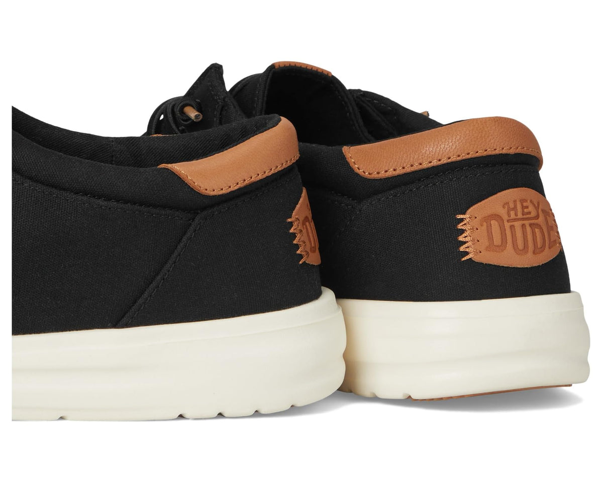Black canvas shoes with tan heel trim and side “HEY DUDE” patch, rear view